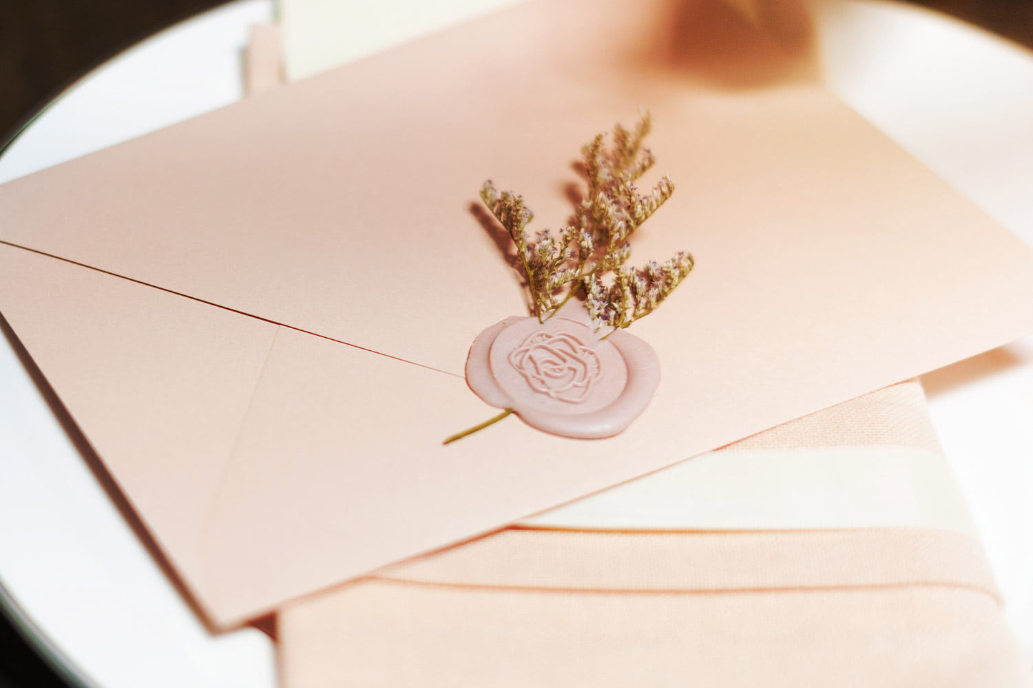 A blush Galentine's envelope sealed with a light pink, rose stamp rests on a dinner napkin and a plate. Dried lavender is pressed into the wax