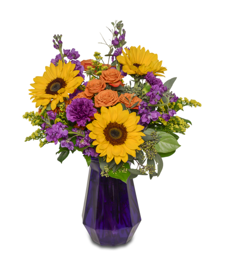 A vibrant floral arrangement in a purple vase with sunflowers, purple stock, purple carnations, orange spray roses, and solidago, hand-arranged with care.
