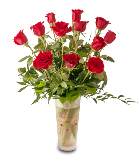 A romantic Valentine’s arrangement of fresh red roses and white baby’s breath arranged in a clear glass vase with heart accents and lush green foliage.