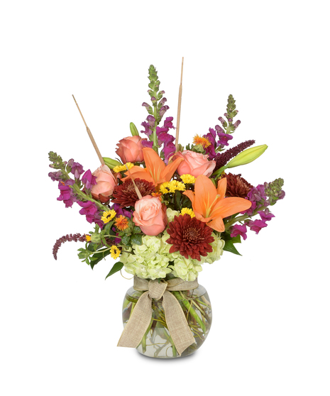 Handcrafted arrangement in recycled glass vase: bright snapdragons, two-tone roses, and orange lilies on a green base; tied with a burlap knot. Deluxe versions enriched.