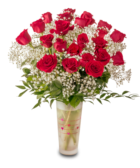 A romantic Valentine’s arrangement of fresh red roses and white baby’s breath arranged in a clear glass vase with heart accents and lush green foliage.