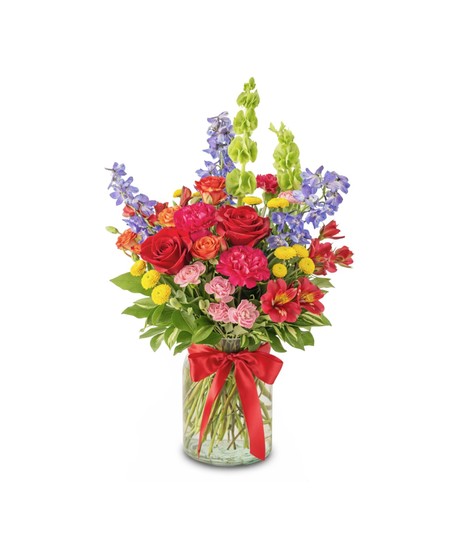 A colorful floral arrangement featuring red roses, hot pink carnations, red alstroemeria, and orange spray roses arranged in a clear glass vase with a red bow.