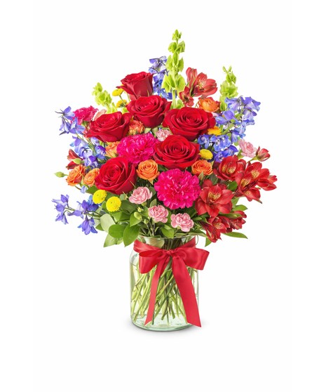 A colorful floral arrangement featuring red roses, hot pink carnations, red alstroemeria, and orange spray roses arranged in a clear glass vase with a red bow.