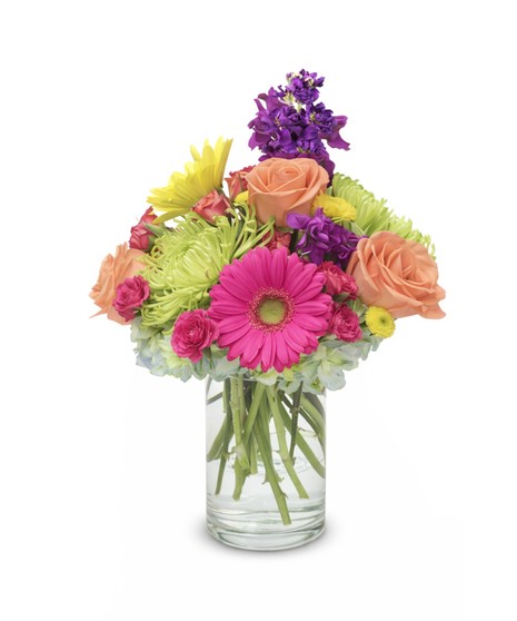 Bright mixed flower arrangement with hot pink, yellow, and orange gerbera daisies, roses, mums, and carnations in a clear glass cylinder vase.