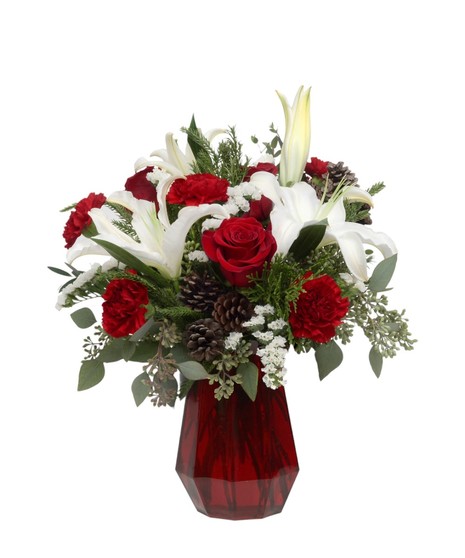 Red and white winter bouquet with lilies, roses, carnations, pinecones, and cedar greenery in a red glass vase arranged for a cozy cabin Christmas feel.