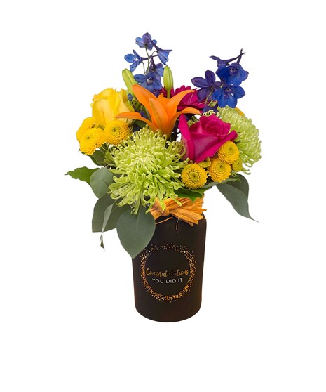 Bright mixed flower arrangement with lilies, roses, green spider mums, and blue delphinium in a black  or white jar that has a graduation message.