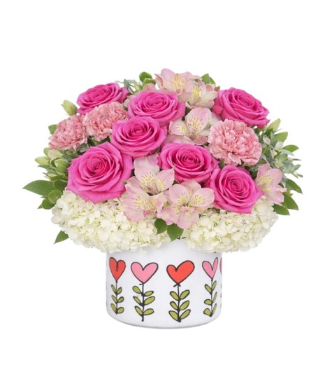 Petite floral arrangement with pink roses, hydrangeas, carnations, and alstroemeria arranged in a white container decorated with colorful heart designs.