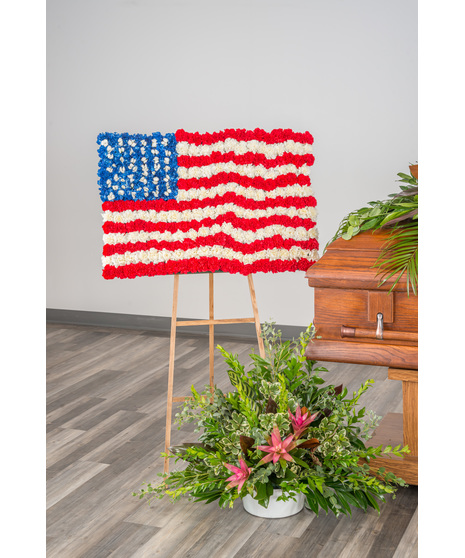 Standing floral arrangement shaped like the American flag in red, white, and blue flowers displayed on a wooden easel place into a Funeral Home setting