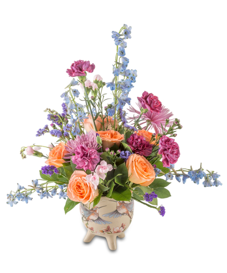 Blue delphinium, coral roses, purple and soft pink flowers arranged with greenery in a decorative hummingbird pot, creating a colorful garden-style spring bouquet.
