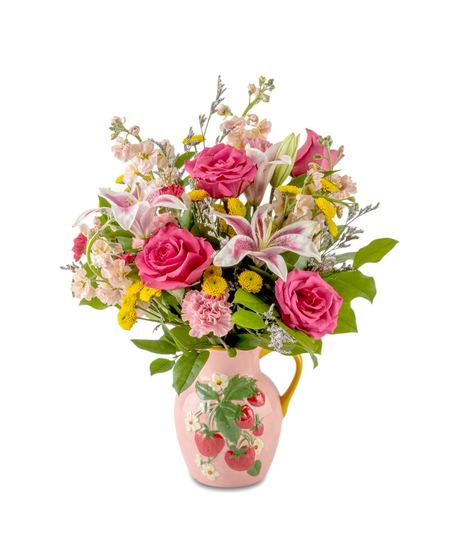 Pink roses, fragrant lilies, stock, and yellow accent flowers arranged in a pink strawberry pitcher vase, creating a cheerful pink and yellow bouquet with lush greenery.