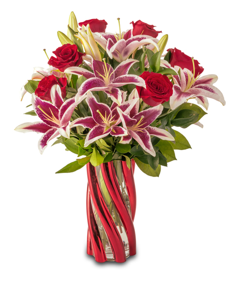 A floral arrangement featuring pink lilies with bright pink centers, red roses, and green foliage arranged in a red glass vase against a white background.
